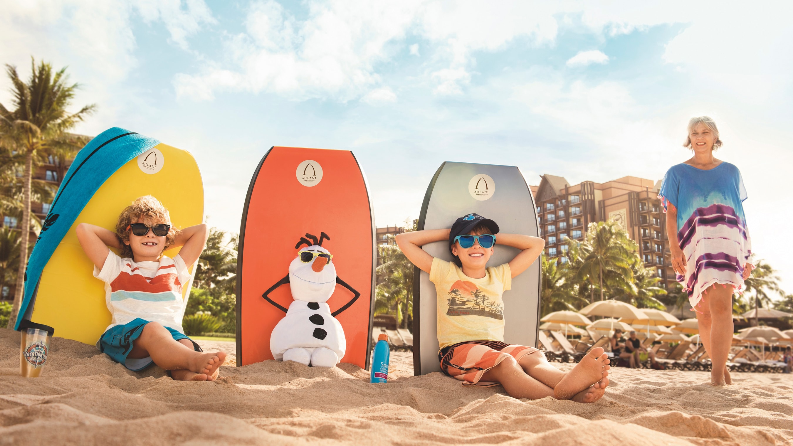 Two boys sit on the beach with Olaf the snowman, while their grandmother stands nearby