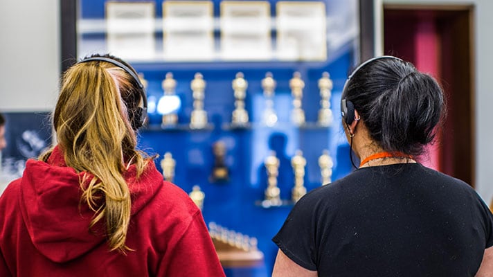 Women wearing headphones, standing in front of a display of trophies